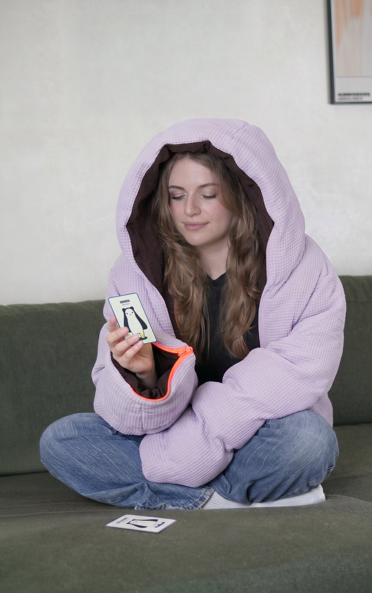 Eine Person sitzt in der fliederfarbenen Kissenjacke im Schneidersitz auf einem Sofa und liest eine Übungskarte, eine weitere Karte liegt daneben.