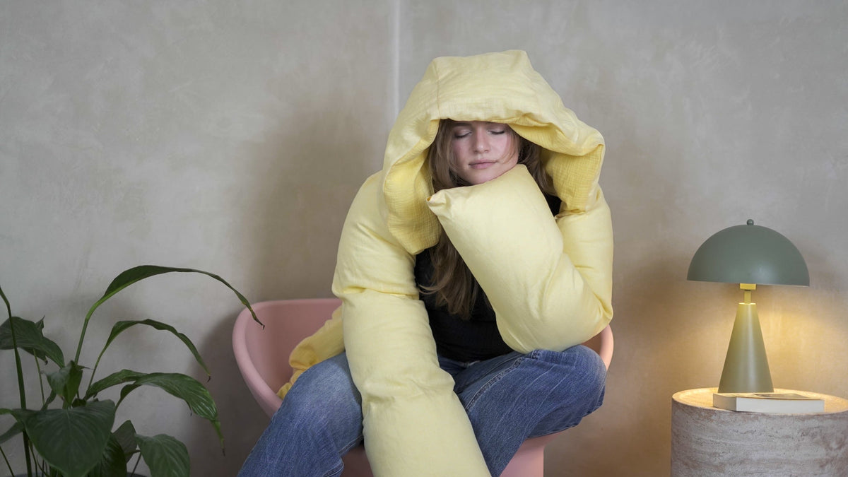 Eine Person in der gelben Kissenjacke Kija sitzt in einem Sessel, die Kapuze über dem Kopf, den Kopf auf den beschwerten Ärmel gelegt, die Augen geschlossen.
